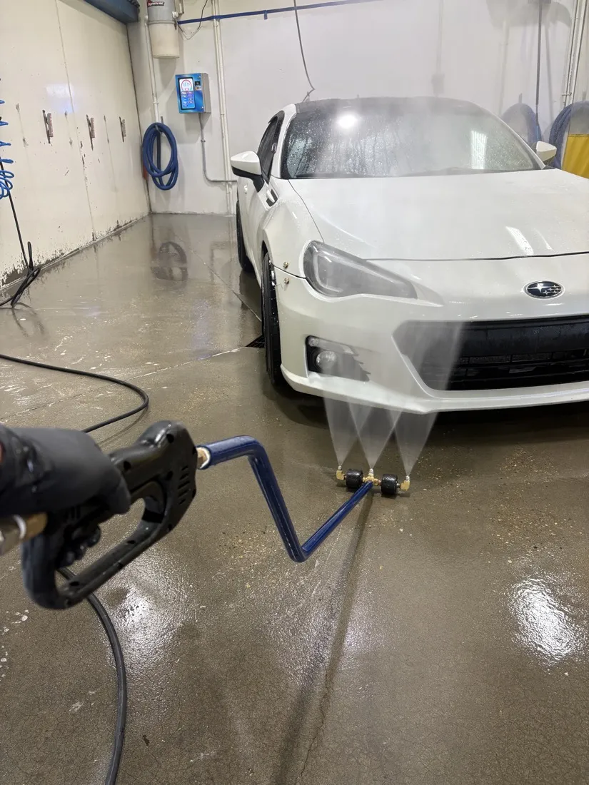 A hand-held undercarriage washer spraying beneath a white car in a self-serve bay